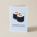 A folded greeting card standing upright on a beige surface. The card has a pale blue front with an illustration of two sushi rolls and the text 'CONGRATULATIONS ON YOUR NEW ROLL!'.