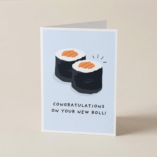 A folded greeting card standing upright on a beige surface. The card has a pale blue front with an illustration of two sushi rolls and the text 'CONGRATULATIONS ON YOUR NEW ROLL!'.