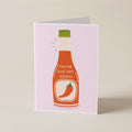 A folded greeting card standing upright on a light beige surface. The card features a pink background with a stylized illustration of a hot sauce bottle in orange-red, with a green cap and the text 'YOU'RE ONE HOT, MAMA!' above a red chili pepper graphic.