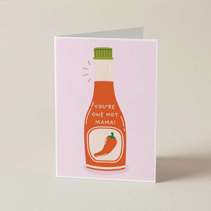A folded greeting card standing upright on a light beige surface. The card features a pink background with a stylized illustration of a hot sauce bottle in orange-red, with a green cap and the text 'YOU'RE ONE HOT, MAMA!' above a red chili pepper graphic.