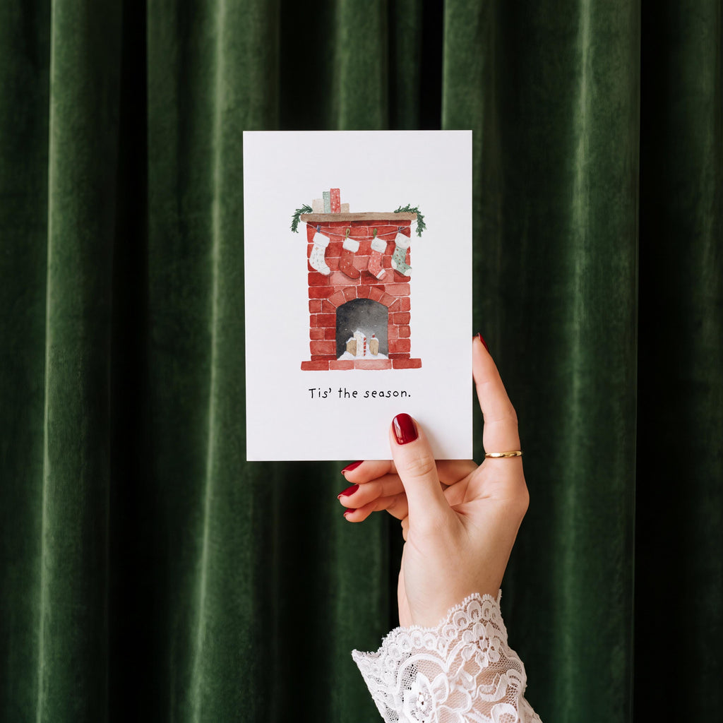A hand with red nail polish and a white lace cuff holds a white greeting card featuring a watercolor illustration of a red brick fireplace with stockings and the text 'Tis the season.' The background is dark green velvet curtains.