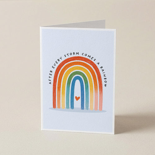 A folded white greeting card standing upright on a beige surface, printed with a rainbow arch, a small red heart, and the text 'AFTER EVERY STORM COMES A RAINbow'.