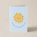 A folded greeting card standing upright on a beige surface. The card has a light blue background with a cute yellow smiling sun illustration in the center and the phrase 'ANOTHER YEAR AROUND THE SUN!' in small black curved text below it.