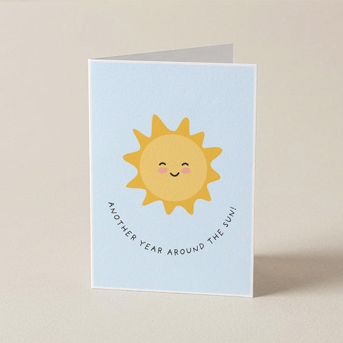 A folded greeting card standing upright on a beige surface. The card has a light blue background with a cute yellow smiling sun illustration in the center and the phrase 'ANOTHER YEAR AROUND THE SUN!' in small black curved text below it.