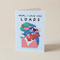 A folded greeting card standing upright on a beige surface. The card has a light blue border and reads 'MUM, I LOVE YOU LOADS' above an illustration of a pile of colorful laundry in a basket.