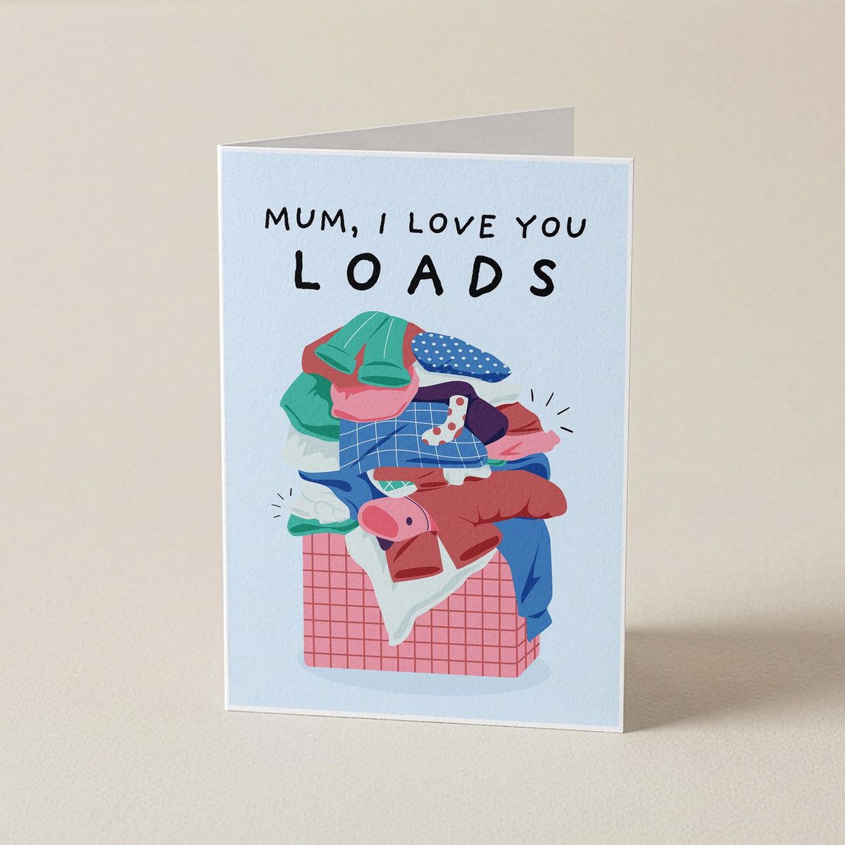 A folded greeting card standing upright on a beige surface. The card has a light blue border and reads 'MUM, I LOVE YOU LOADS' above an illustration of a pile of colorful laundry in a basket.