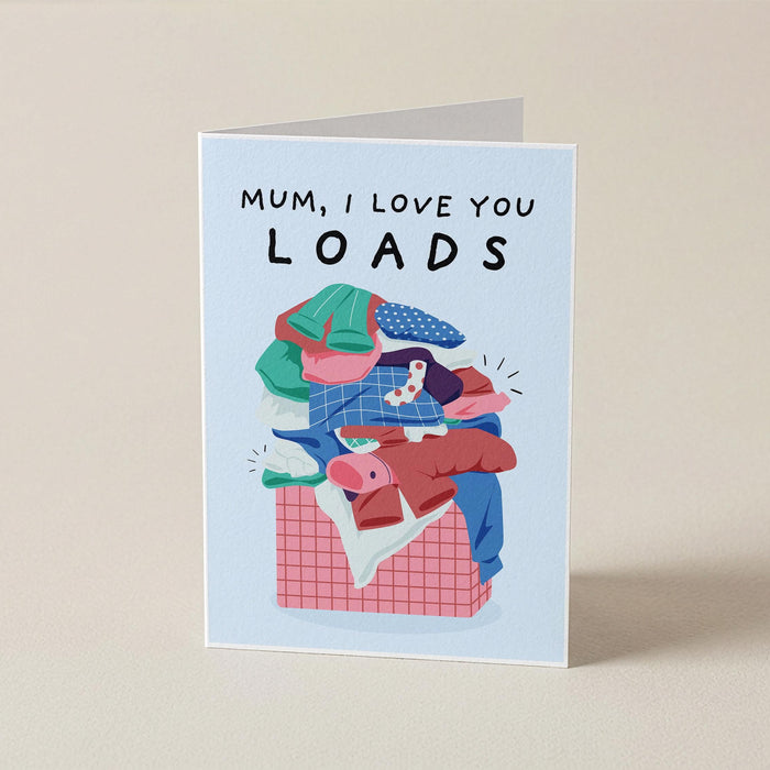 A folded greeting card standing upright on a beige surface. The card has a light blue border and reads 'MUM, I LOVE YOU LOADS' above an illustration of a pile of colorful laundry in a basket.