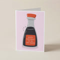 A folded greeting card standing upright on a light beige surface. The card features a minimalist illustration of a soy sauce bottle with a red cap and a red label that reads 'HAPPY BIRTHDAY TO MY SOY-MATE' in white text, set against a pale pink background.
