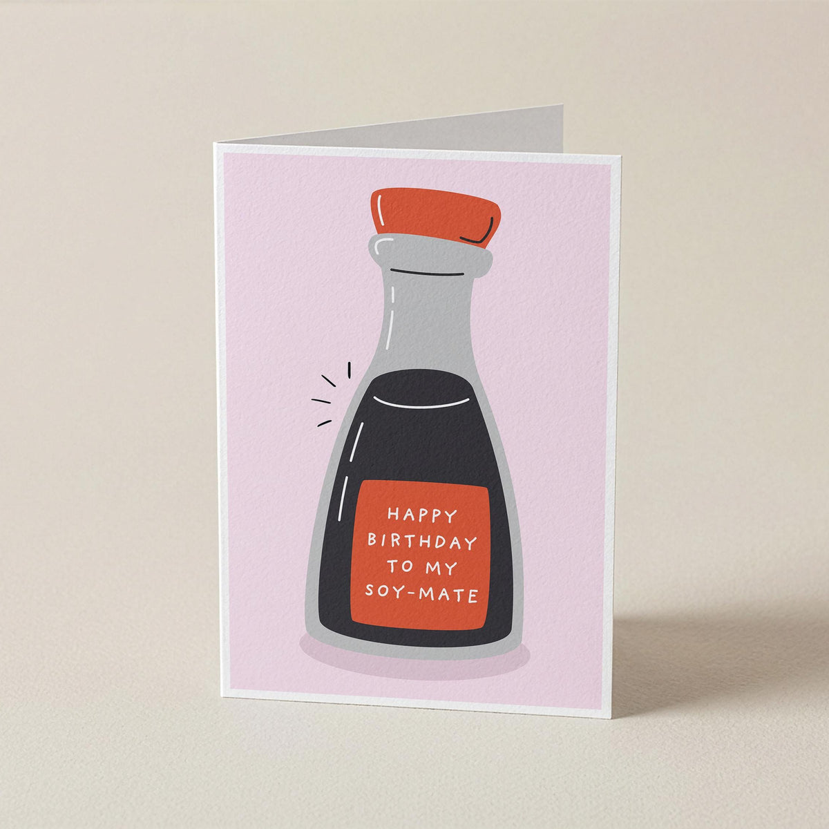 A folded greeting card standing upright on a light beige surface. The card features a minimalist illustration of a soy sauce bottle with a red cap and a red label that reads 'HAPPY BIRTHDAY TO MY SOY-MATE' in white text, set against a pale pink background.