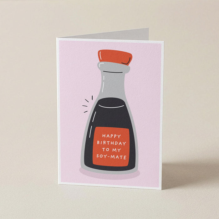 A folded greeting card standing upright on a light beige surface. The card features a minimalist illustration of a soy sauce bottle with a red cap and a red label that reads 'HAPPY BIRTHDAY TO MY SOY-MATE' in white text, set against a pale pink background.