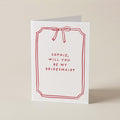 A white greeting card with a red border, red bow illustration, and the text 'SOPHIE, WILL YOU BE MY BRIDESMAID?' on a warm beige background.