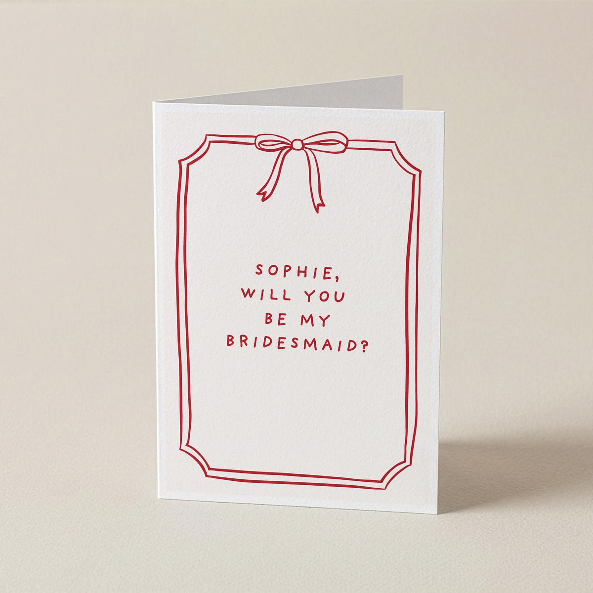 A white greeting card with a red border, red bow illustration, and the text 'SOPHIE, WILL YOU BE MY BRIDESMAID?' on a warm beige background.