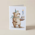 A folded greeting card featuring a watercolor illustration of a teddy bear wearing a party hat and bow, holding a birthday cake with a number 1 candle, set against a plain beige background.