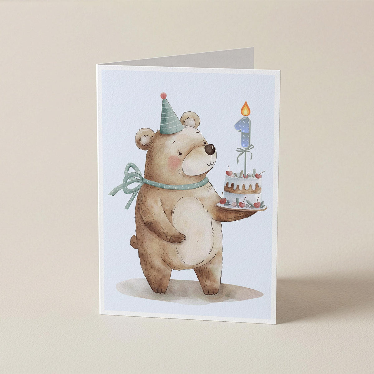 A folded greeting card featuring a watercolor illustration of a teddy bear wearing a party hat and bow, holding a birthday cake with a number 1 candle, set against a plain beige background.