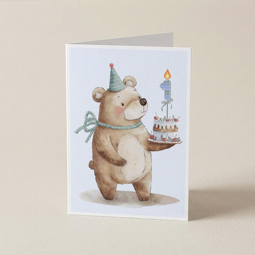 A folded greeting card featuring a watercolor illustration of a teddy bear wearing a party hat and bow, holding a birthday cake with a number 1 candle, set against a plain beige background.