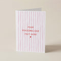 A vertical greeting card with pink and white vertical stripes, featuring the text 'YOUR PERSONALISED TEXT HERE' and a small heart in red, standing upright on a neutral beige background.