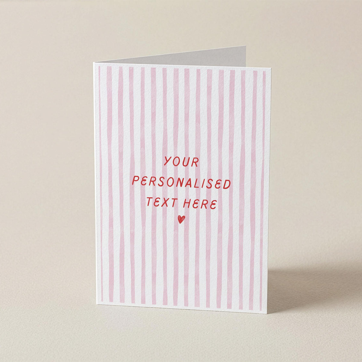 A vertical greeting card with pink and white vertical stripes, featuring the text 'YOUR PERSONALISED TEXT HERE' and a small heart in red, standing upright on a neutral beige background.