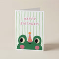 A folded greeting card standing upright on a light beige surface. The card features a green cartoon frog peeking up from the bottom with pink cheeks, a pink party hat, and the words 'HAPPY BIRTHDAY!' in pink against a white background with pale green vertical stripes.