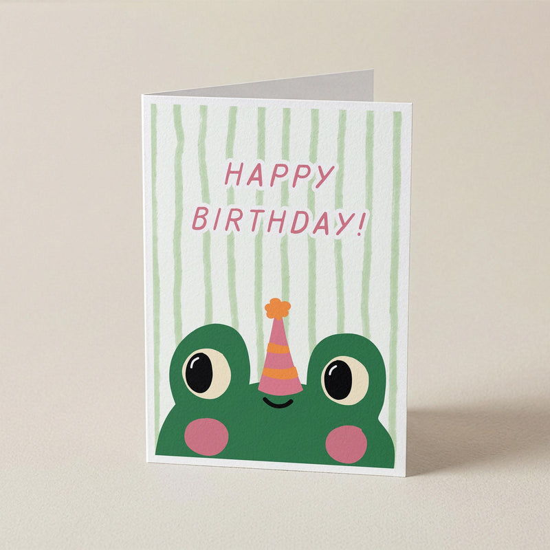 A folded greeting card standing upright on a light beige surface. The card features a green cartoon frog peeking up from the bottom with pink cheeks, a pink party hat, and the words 'HAPPY BIRTHDAY!' in pink against a white background with pale green vertical stripes.