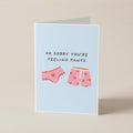 A folded greeting card standing upright on a beige surface. The card has a light blue background and features the text 'SO SORRY YOU'RE FEELING PANTS' above two pink underwear illustrations with small red hearts.