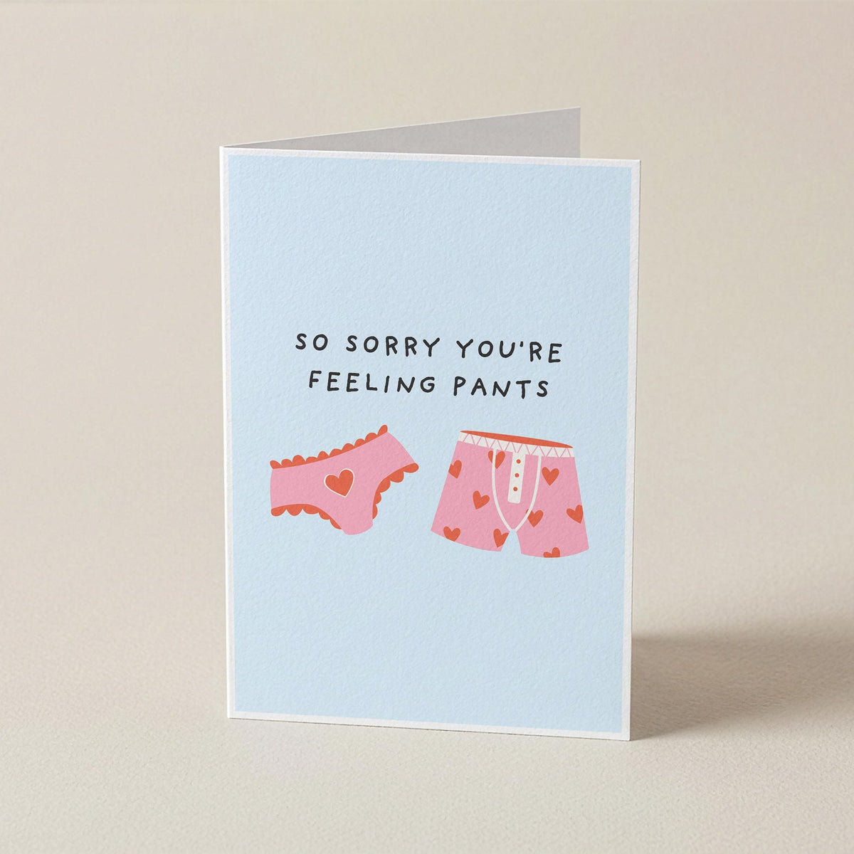 A folded greeting card standing upright on a beige surface. The card has a light blue background and features the text 'SO SORRY YOU'RE FEELING PANTS' above two pink underwear illustrations with small red hearts.