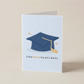 A folded graduation card standing upright on a light beige surface. The front of the card is pale blue with a navy blue graduation cap illustration, a gold tassel, small gold stars, and the word 'CONGRADULATIONS!' printed below.