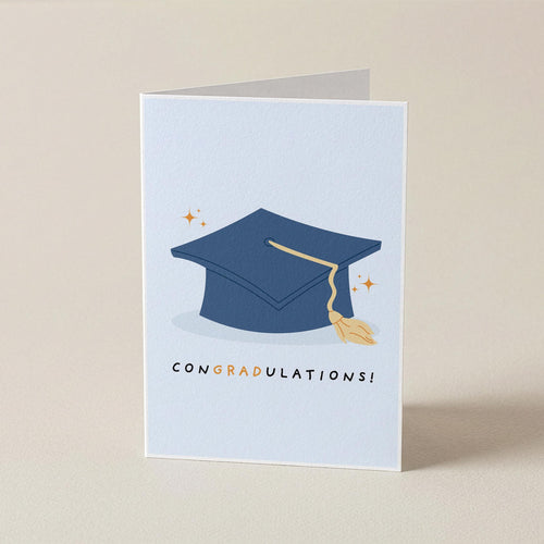 A folded graduation card standing upright on a light beige surface. The front of the card is pale blue with a navy blue graduation cap illustration, a gold tassel, small gold stars, and the word 'CONGRADULATIONS!' printed below.