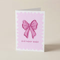 A folded greeting card standing upright on a light beige surface. The front of the card has a white border, a pink scalloped square background, a pink bow illustration in the center, and the words 'BIRTHDAY BABE' printed below.