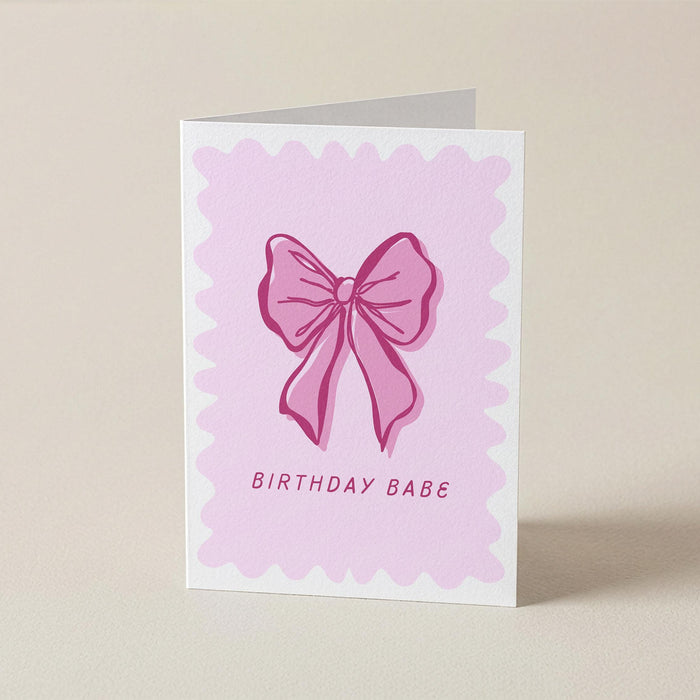 A folded greeting card standing upright on a light beige surface. The front of the card has a white border, a pink scalloped square background, a pink bow illustration in the center, and the words 'BIRTHDAY BABE' printed below.