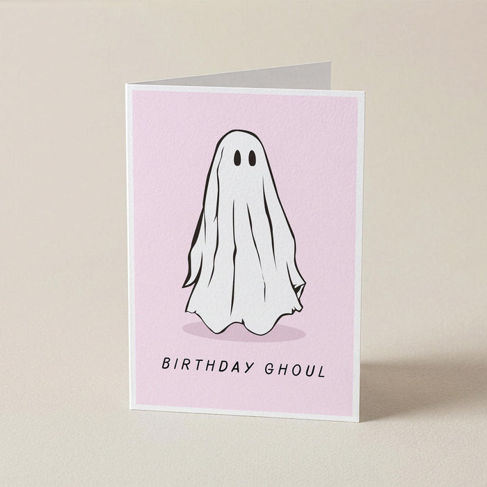 A folded greeting card standing upright on a light beige surface. The card has a pale pink front with a simple black-and-white ghost illustration and the words 'BIRTHDAY GHOUL' printed near the bottom.