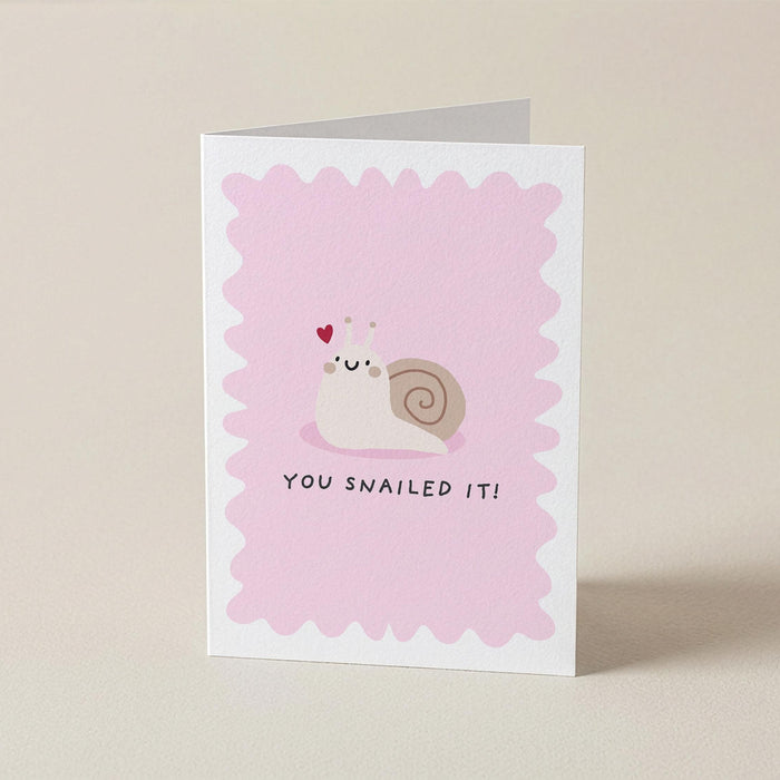 A folded greeting card standing upright on a light beige surface. The front of the card has a pink scalloped border, a small cartoon snail with a heart on its forehead, and the text 'YOU SNAILED IT!' below.
