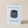 A folded greeting card standing upright on a light beige surface. The card has a white background and features a black-and-gray illustration of an air fryer with the text 'ONE MINUTE YOU'RE YOUNG, THE NEXT YOU'RE EXCITED ABOUT AN AIRFRYER!'