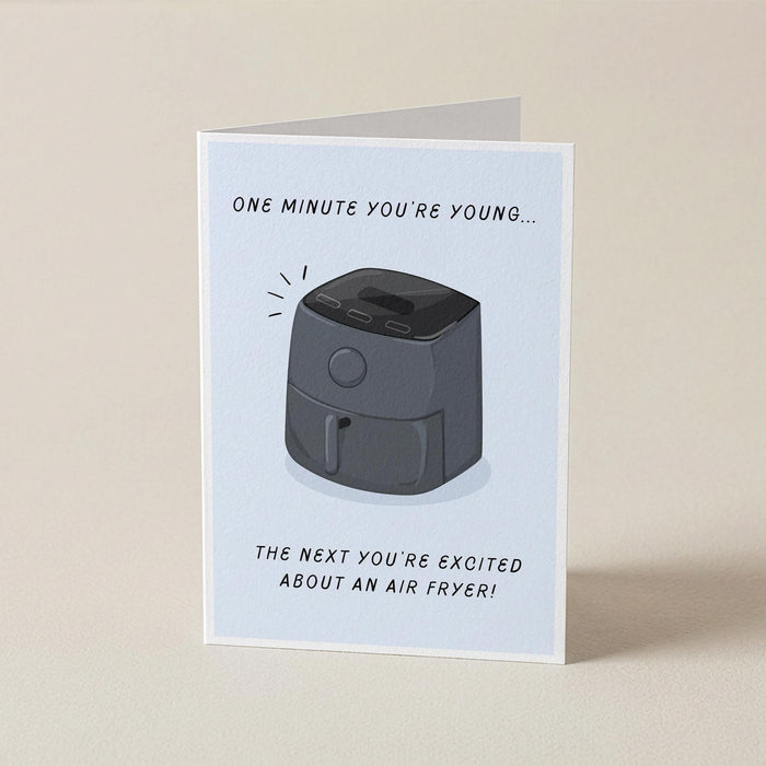 A folded greeting card standing upright on a light beige surface. The card has a white background and features a black-and-gray illustration of an air fryer with the text 'ONE MINUTE YOU'RE YOUNG, THE NEXT YOU'RE EXCITED ABOUT AN AIRFRYER!'