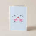 A folded greeting card standing upright on a light beige surface. The card has a pale blue front with a printed illustration of two pink birds facing each other under the text 'you pair of lovebirds!'.