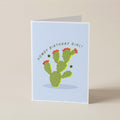 A folded greeting card standing upright on a light beige surface. The card has a pale blue front with a green cactus illustration, small red flowers, and the text 'HOWDY BIRTHDAY GIRL!' curved above the cactus.