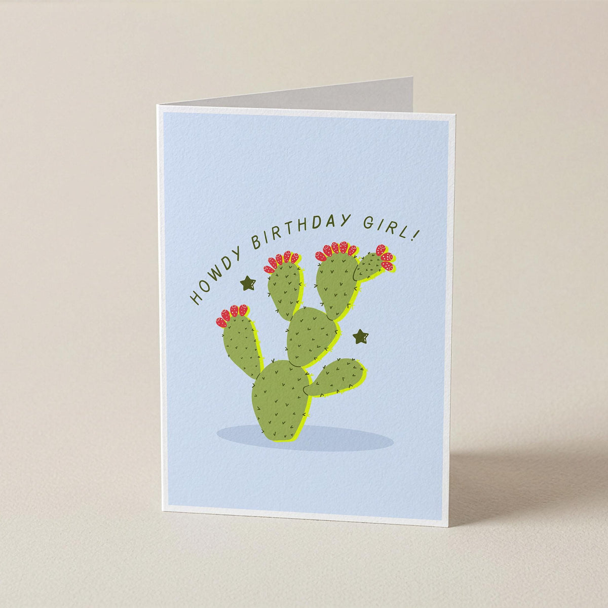 A folded greeting card standing upright on a light beige surface. The card has a pale blue front with a green cactus illustration, small red flowers, and the text 'HOWDY BIRTHDAY GIRL!' curved above the cactus.