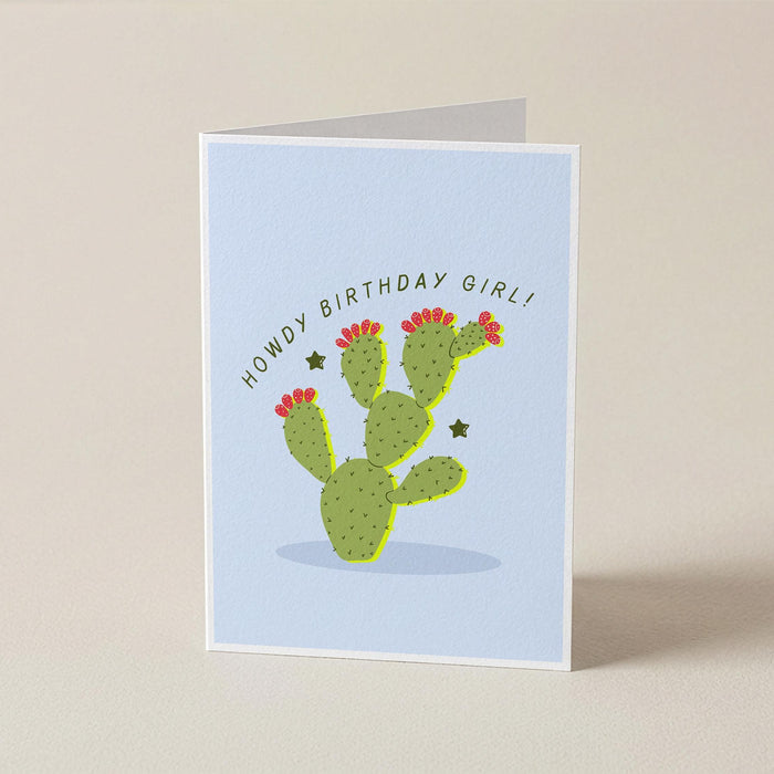 A folded greeting card standing upright on a light beige surface. The card has a pale blue front with a green cactus illustration, small red flowers, and the text 'HOWDY BIRTHDAY GIRL!' curved above the cactus.