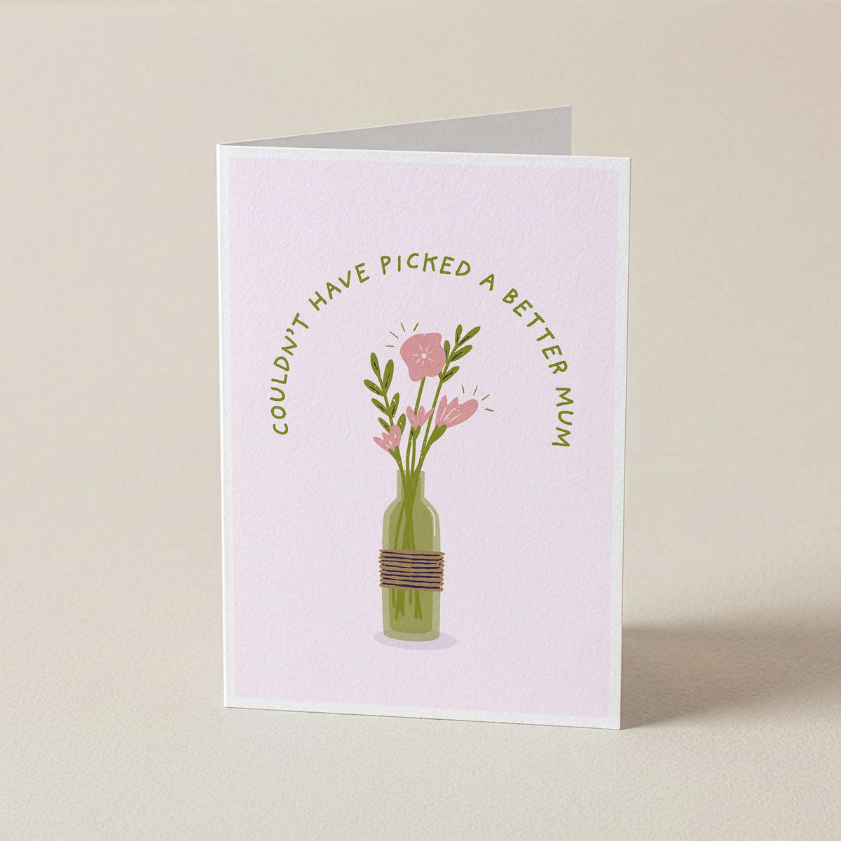 A folded greeting card standing upright on a light beige surface. The card has a pale pink background with an illustration of pink flowers in a green bottle vase, and curved text that reads, 'COULDN'T HAVE PICKED A BETTER MOM.