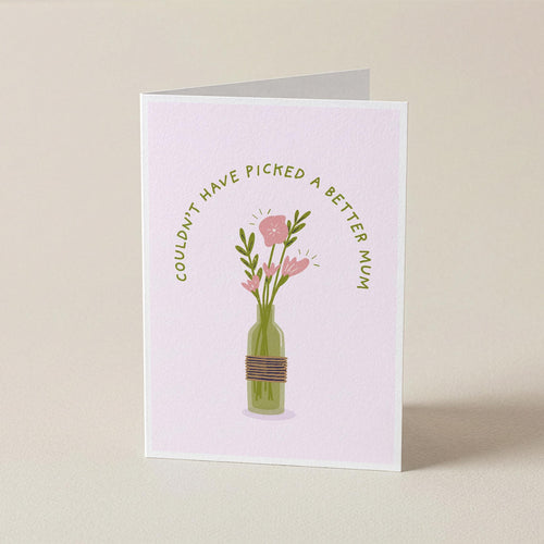 A folded greeting card standing upright on a light beige surface. The card has a pale pink background with an illustration of pink flowers in a green bottle vase, and curved text that reads, 'COULDN'T HAVE PICKED A BETTER MOM.