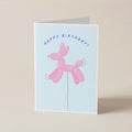 A folded greeting card standing upright on a light beige surface. The card has a pale blue front with the words “HAPPY BIRTHDAY!” above a pink balloon-dog illustration.