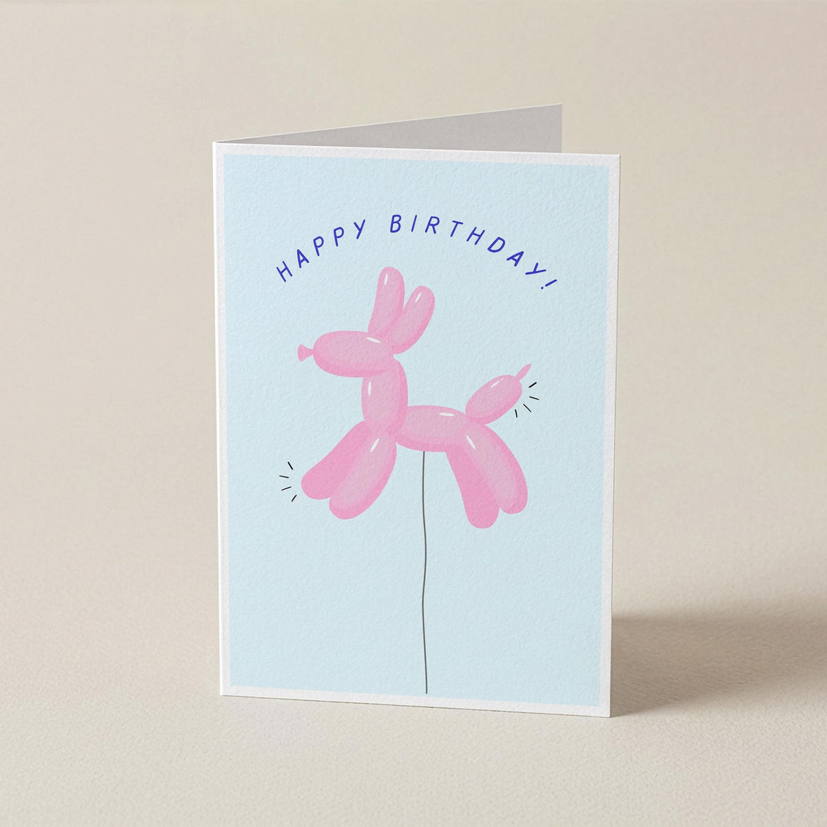 A folded greeting card standing upright on a light beige surface. The card has a pale blue front with the words “HAPPY BIRTHDAY!” above a pink balloon-dog illustration.