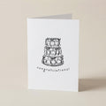 A white greeting card standing upright on a light beige surface, printed with a black line drawing of a three-tiered cake and the word 'congratulations!' below it.