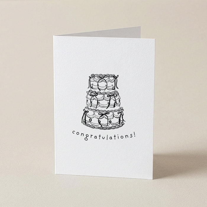 A white greeting card standing upright on a light beige surface, printed with a black line drawing of a three-tiered cake and the word 'congratulations!' below it.