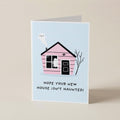 A folded greeting card standing upright on a light beige surface. The card features a light blue background with a pink house illustration, a white ghost, and the text 'HOPE YOUR NEW HOUSE ISN'T HAUNTED!' in black lettering.