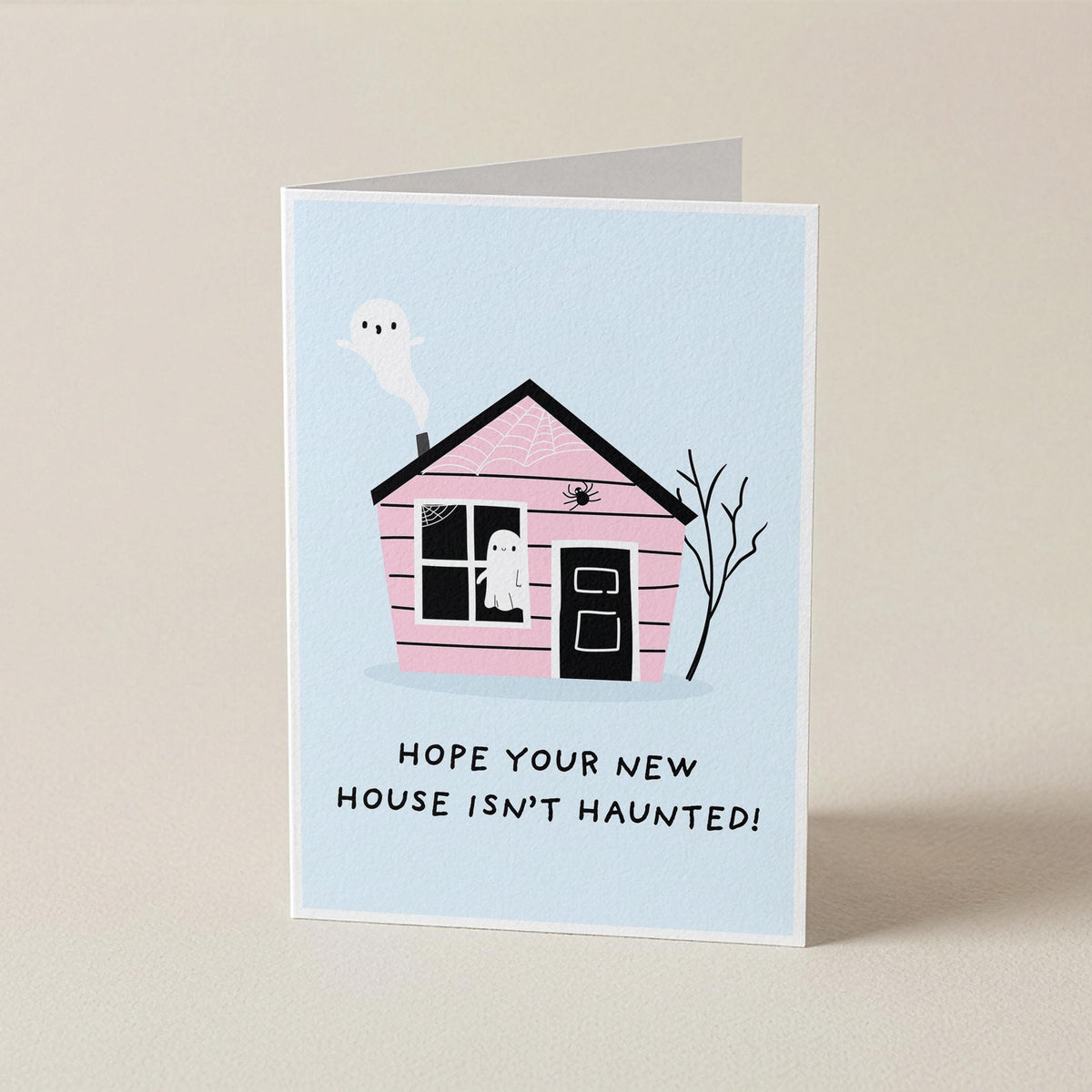 A folded greeting card standing upright on a light beige surface. The card features a light blue background with a pink house illustration, a white ghost, and the text 'HOPE YOUR NEW HOUSE ISN'T HAUNTED!' in black lettering.