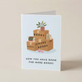 A folded greeting card standing upright on a beige surface. The card features an illustration of stacked cardboard boxes labeled 'BOOKS' and 'MORE BOOKS' with a small potted plant on top, and the text 'NOW YOU HAVE ROOM FOR MORE BOOKS!' below.