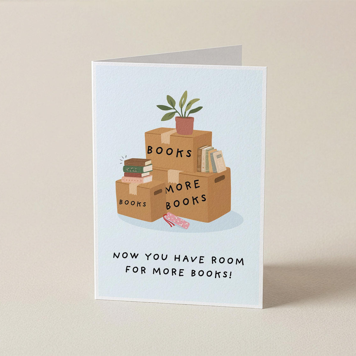 A folded greeting card standing upright on a beige surface. The card features an illustration of stacked cardboard boxes labeled 'BOOKS' and 'MORE BOOKS' with a small potted plant on top, and the text 'NOW YOU HAVE ROOM FOR MORE BOOKS!' below.