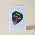 A folded greeting card standing upright on a light beige surface. The card has a pale blue front with a black guitar-pick illustration containing the words 'YOU ROCK!' in white, plus small orange flames at the bottom of the pick.