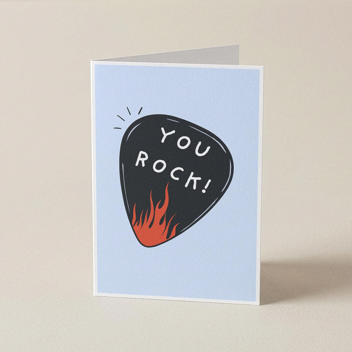 A folded greeting card standing upright on a light beige surface. The card has a pale blue front with a black guitar-pick illustration containing the words 'YOU ROCK!' in white, plus small orange flames at the bottom of the pick.