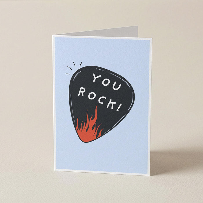 A folded greeting card standing upright on a light beige surface. The card has a pale blue front with a black guitar-pick illustration containing the words 'YOU ROCK!' in white, plus small orange flames at the bottom of the pick.